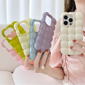 May include: A collection of colorful phone cases in shades of pink, green, yellow, blue, and purple. Each case has a unique, textured design. The cases are held up in front of a person.