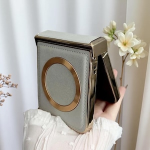May include: A gray phone case with a gold-colored circular accent and trim. The case is open, revealing a dark interior. The case is held in a hand, with a white floral arrangement in the background.