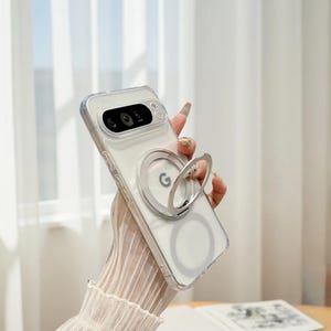 Può includere: Custodia trasparente per telefono con supporto ad anello argentato. La custodia ha un retro trasparente e un anello argentato con la lettera "G". La custodia del telefono è progettata per adattarsi a un modello di telefono specifico. La custodia è tenuta da una persona.