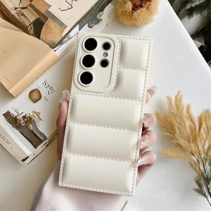 Puede incluir: Funda de teléfono de color crema con un diseño acolchado. La funda tiene un recorte para la cámara con múltiples lentes y un borde cosido. La funda del teléfono la sostiene una persona.