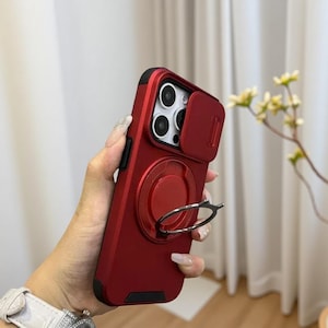 Op de afbeelding: Rode telefoonhoes met een cameralensafdekking en een ringhouder. De hoes heeft een zwarte rand en een cirkelvormige ringhouder aan de achterkant. De cameralensafdekking is ook rood en schuift open en dicht.