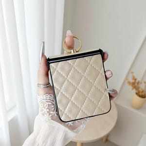 May include: A beige quilted phone case with a black border and a gold ring holder. The case has a diamond pattern and a smooth, leather-like texture. The ring is attached to the top of the case.