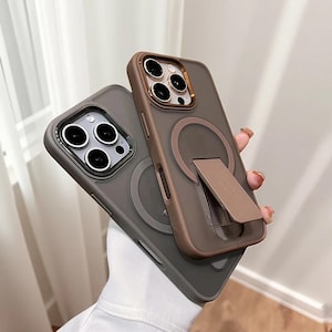 Puede incluir: Dos fundas para teléfono, una gris y otra marrón, están apiladas. La funda gris tiene un anillo de cámara plateado y un círculo magnético en la parte trasera. La funda marrón tiene un anillo de cámara dorado, un círculo magnético y un soporte.