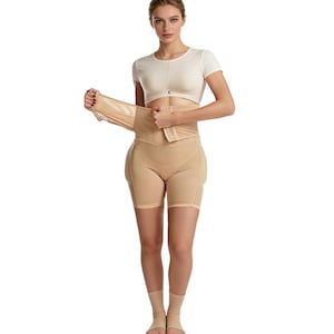 May include: A beige body shaper garment, featuring a high-waisted design with shorts and a waist cincher. The garment has a hook-and-eye closure and is made of a smooth, seamless material. The model is wearing a white crop top.