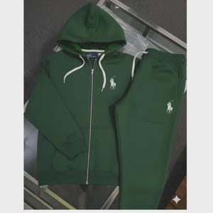 May include: A dark green tracksuit set featuring a hooded zip-up jacket and matching sweatpants. The jacket and pants have a white logo on the chest and leg. The jacket has white drawstrings and a full zipper. The set is made of a soft material.