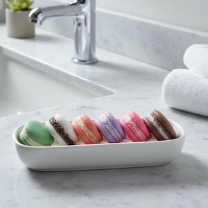 Op de afbeelding: Een wit keramisch dienblad bevat zes kleurrijke macarons. De macarons zijn groen, wit met bruine vulling, oranje, lavendel, roze en bruin. De lade staat op een marmeren oppervlak in de buurt van een wastafel en kraan.