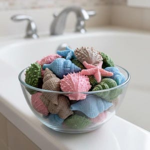 May include: A glass bowl filled with colourful seashell-shaped soaps. The soaps are in shades of pink, blue, green, and tan, including starfish and various shell designs. The bowl is on a white surface, with a blurred bathroom sink in the background.