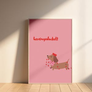 Könnte beinhalten: Gerahmter rosa Druck mit einem Cartoon-Dackel mit roter Schleife und herzförmiger Girlande. Der deutsche Ausdruck „hereingedackelt“ steht in Rot oben. Das Kunstwerk wird auf einem Holzboden ausgestellt.