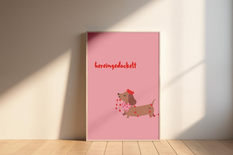 Könnte beinhalten: Gerahmter rosa Druck mit einem Cartoon-Dackel mit roter Schleife und herzförmiger Girlande. Der deutsche Ausdruck „hereingedackelt“ steht in Rot oben. Das Kunstwerk wird auf einem Holzboden ausgestellt.
