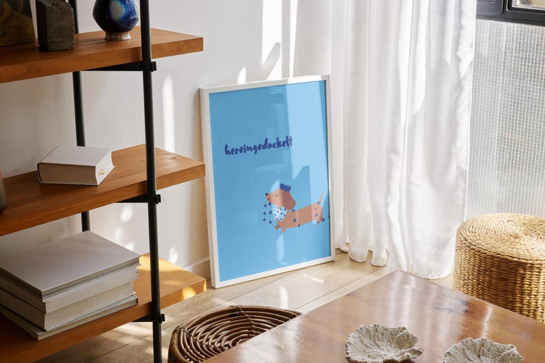 K&ouml;nnte beinhalten: Ein gerahmter Kunstdruck mit einer Dackel-Illustration auf hellblauem Hintergrund. Der Dackel tr&auml;gt einen Hut und einen gepunkteten Schal. Der Text "hereingedackelt" steht &uuml;ber dem Hund. Der Druck wird auf einem Holzboden pr&auml;sentiert.