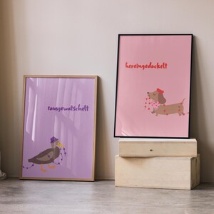 K&ouml;nnte beinhalten: Zwei gerahmte Kunstdrucke. Einer zeigt eine Ente mit lila Hut und dem Text "rausgewatschelt". Der andere Druck zeigt einen Dackel mit rotem Hut und dem Text "hereingedackelt". Beide Drucke sind im Cartoon-Stil gehalten.