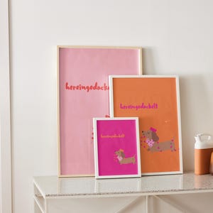 May include: Three framed art prints with dachshund illustrations and the word "hereingedackt" in a playful font. The prints are in pink, orange, and fuchsia, with white and natural wood frames. The artwork is displayed on a white table.