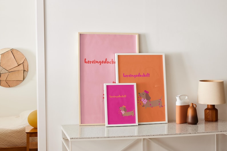 May include: Three framed art prints with dachshund illustrations and the word "hereingedackt" in a playful font. The prints are in pink, orange, and fuchsia, with white and natural wood frames. The artwork is displayed on a white table.