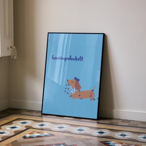 K&ouml;nnte beinhalten: Gerahmter Kunstdruck mit einem Cartoon-Dackel mit blauer Schleife und gepunktetem Schal auf hellblauem Hintergrund. Der Druck zeigt das Wort "heringgedackelt" in Schreibschrift.
