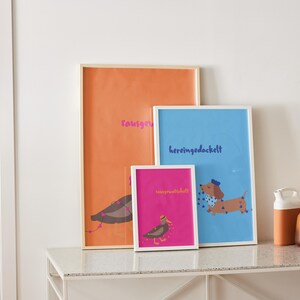 K&ouml;nnte beinhalten: Drei gerahmte Kunstdrucke mit Illustrationen eines Vogels und eines Dackelhundes. Die Drucke sind in Orange, Blau und Pink gehalten und haben wei&szlig;e Rahmen. Der orangefarbene Druck tr&auml;gt das Wort "rausgeknallt" in Pink. Der blaue Druck tr&auml;gt das Wort "hereingedockt" in Blau.