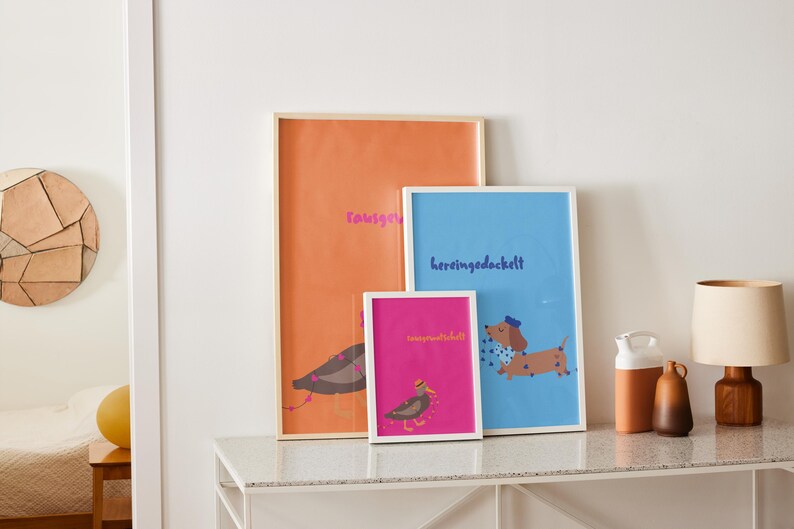 K&ouml;nnte beinhalten: Drei gerahmte Kunstdrucke mit Illustrationen eines Vogels und eines Dackelhundes. Die Drucke sind in Orange, Blau und Pink gehalten und haben wei&szlig;e Rahmen. Der orangefarbene Druck tr&auml;gt das Wort "rausgeknallt" in Pink. Der blaue Druck tr&auml;gt das Wort "hereingedockt" in Blau.