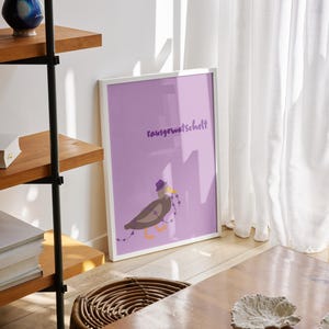 K&ouml;nnte beinhalten: Ein gerahmter Kunstdruck mit einem lavendelfarbenen Hintergrund, der einen Cartoonvogel mit Hut und Lichterkette zeigt. Der Druck enth&auml;lt das Wort "rausgewatschelt". Das Kunstwerk wird auf einem Holzboden mit einem Weidenkorb und einem Holztisch ausgestellt.