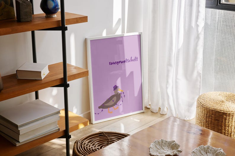 K&ouml;nnte beinhalten: Ein gerahmter Kunstdruck mit einem lavendelfarbenen Hintergrund, der einen Cartoonvogel mit Hut und Lichterkette zeigt. Der Druck enth&auml;lt das Wort "rausgewatschelt". Das Kunstwerk wird auf einem Holzboden mit einem Weidenkorb und einem Holztisch ausgestellt.
