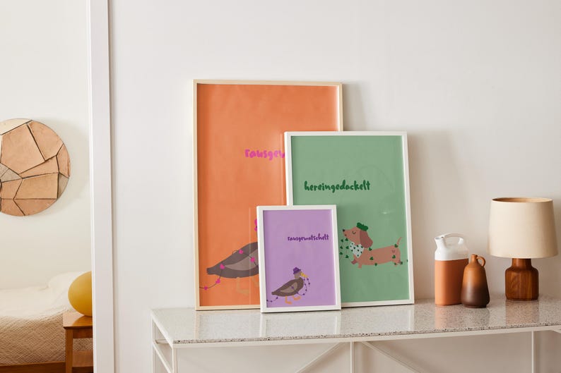 K&ouml;nnte beinhalten: Drei gerahmte Kunstdrucke mit Illustrationen und Text. Der gr&ouml;&szlig;te ist orange mit einem Vogel und rosa Text. Der mittlere Druck ist gr&uuml;n mit einem Dackel und gr&uuml;nem Text. Der kleinste ist lila mit einem Vogel und Text.