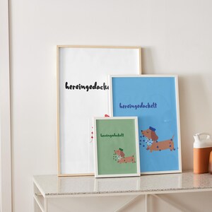 K&ouml;nnte beinhalten: Drei gerahmte Kunstdrucke mit dem Wort "hereingedackelt" in schwarzer Schrift. Die Drucke zeigen eine Dackel-Illustration. Ein Druck ist wei&szlig;, einer blau und einer gr&uuml;n. Die Drucke werden auf einem wei&szlig;en Tisch ausgestellt.