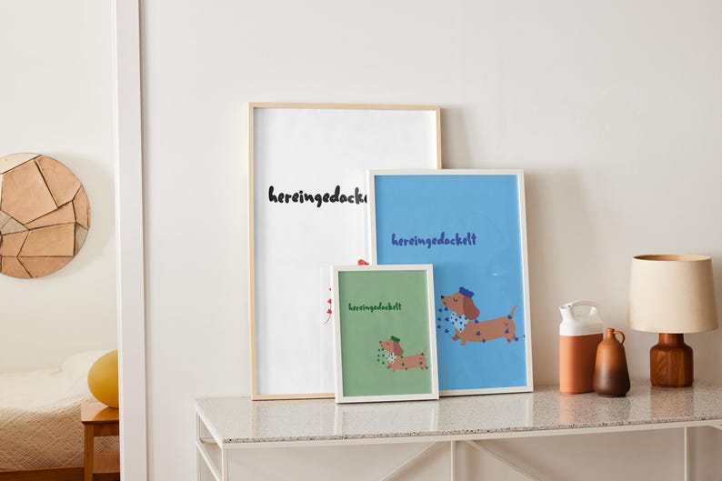 K&ouml;nnte beinhalten: Drei gerahmte Kunstdrucke mit dem Wort "hereingedackelt" in schwarzer Schrift. Die Drucke zeigen eine Dackel-Illustration. Ein Druck ist wei&szlig;, einer blau und einer gr&uuml;n. Die Drucke werden auf einem wei&szlig;en Tisch ausgestellt.