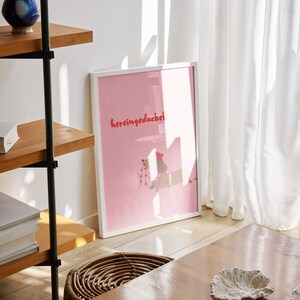 May include: A framed pink art print with the text "hereingedacke" in red lettering. The print is leaning against a white wall. A wooden shelf with books and decorative items is to the left. A woven basket and a wooden table are in the foreground.
