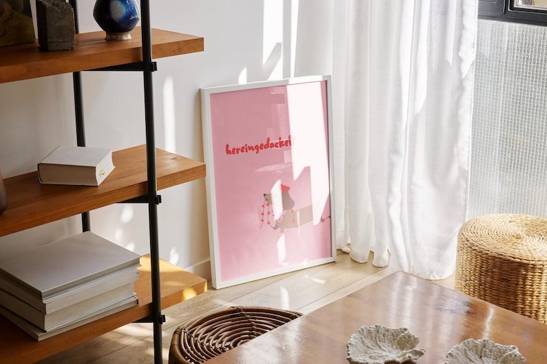 May include: A framed pink art print with the text "hereingedacke" in red lettering. The print is leaning against a white wall. A wooden shelf with books and decorative items is to the left. A woven basket and a wooden table are in the foreground.