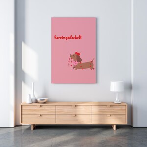 May include: A pink canvas print featuring a cartoon dachshund with a red bow and a scarf adorned with heart shapes. The dachshund is carrying a string of hearts. The text "hereingedackelt" is written in red.