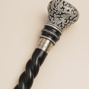 Może przedstawiać: Ozdobna laska do chodzenia ze srebrnym, szczegółowym kwiatowym wzorem na uchwycie. Uchwyt spoczywa na czarnym, skręconym trzonku. Laska posiada srebrne akcenty i ozdobny kołnierz.