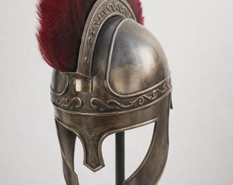 May include: A bronze-colored Roman helmet with a red plume. The helmet features intricate decorative patterns along the brow and sides. The helmet is displayed on a black stand, showcasing its historical design.