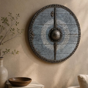 Op de afbeelding: Een rond houten schild met een verweerde blauwe afwerking en horizontale plankenstructuur. Het schild is versierd met een centrale metalen knop, een verticale kruisvormige metalen versteviging en een wit vlechtwerkpatroon.
