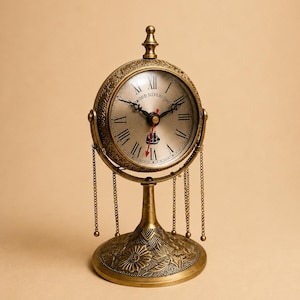 May include: A brass-toned table clock featuring a circular face with Roman numerals, a ship graphic, and the text "WOOD SILVERCORD". The clock is mounted on a pedestal base with floral engravings and is adorned with delicate hanging metal chains.