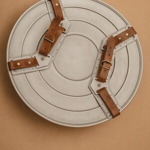 May include: A grey, circular shield with brown leather straps and metal detailing. The shield features concentric circular grooves and is designed with two sets of straps for arm attachment. The straps are riveted and have adjustable buckles.