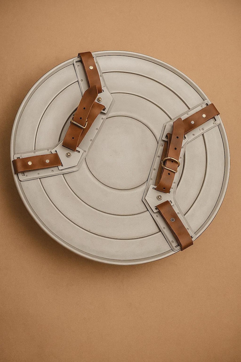 May include: A grey, circular shield with brown leather straps and metal detailing. The shield features concentric circular grooves and is designed with two sets of straps for arm attachment. The straps are riveted and have adjustable buckles.