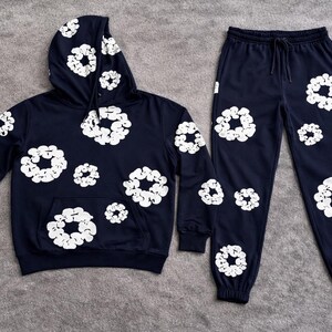 May include: Navy blue hoodie and sweatpants set. The hoodie has a front pocket and drawstring hood. Both pieces are adorned with a white floral pattern. The sweatpants have an elastic waistband and cuffs. The set is displayed on a gray surface.