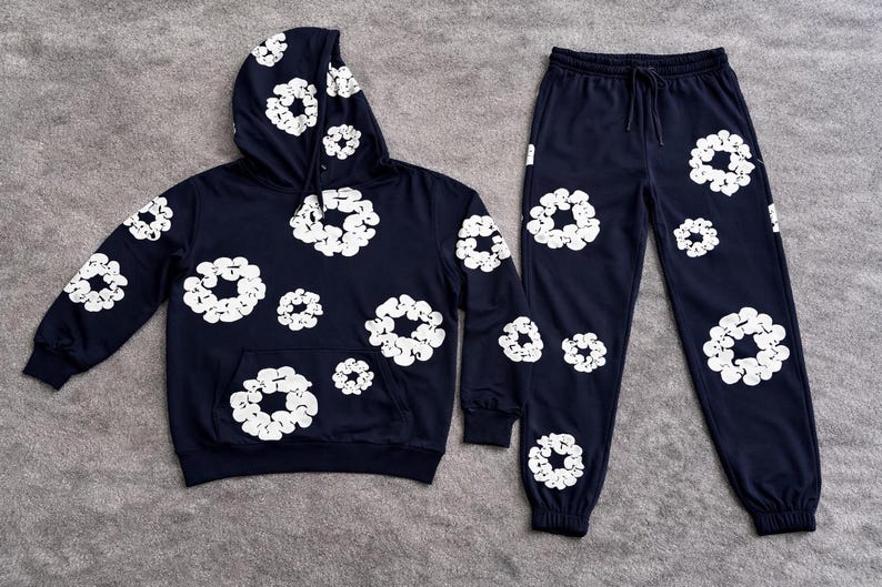 May include: Navy blue hoodie and sweatpants set. The hoodie has a front pocket and drawstring hood. Both pieces are adorned with a white floral pattern. The sweatpants have an elastic waistband and cuffs. The set is displayed on a gray surface.