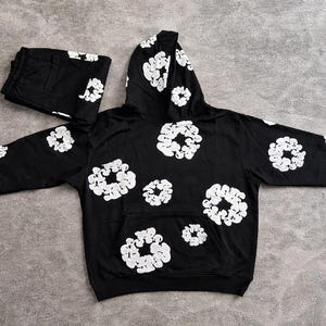 May include: Black hoodie and matching sweatpants set. The set features a repeating white floral pattern. The hoodie has a front pocket and a hood. The sweatpants are folded and placed next to the hoodie.