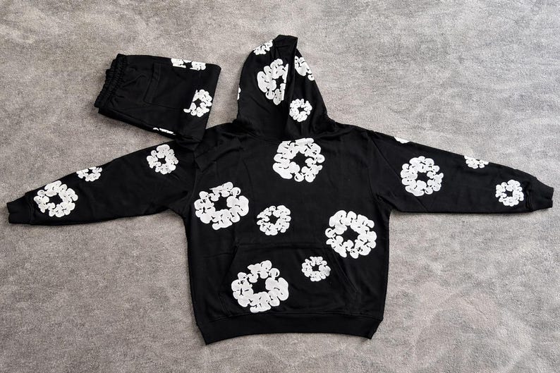 May include: Black hoodie and matching sweatpants set. The set features a repeating white floral pattern. The hoodie has a front pocket and a hood. The sweatpants are folded and placed next to the hoodie.