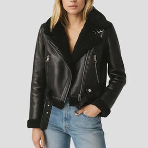 May include: Black leather jacket with a shearling collar and lining. The jacket features a classic asymmetrical zipper closure, multiple zippered pockets, and a belt detail at the waist. The sleeves have shearling cuffs. The jacket is worn over blue jeans.