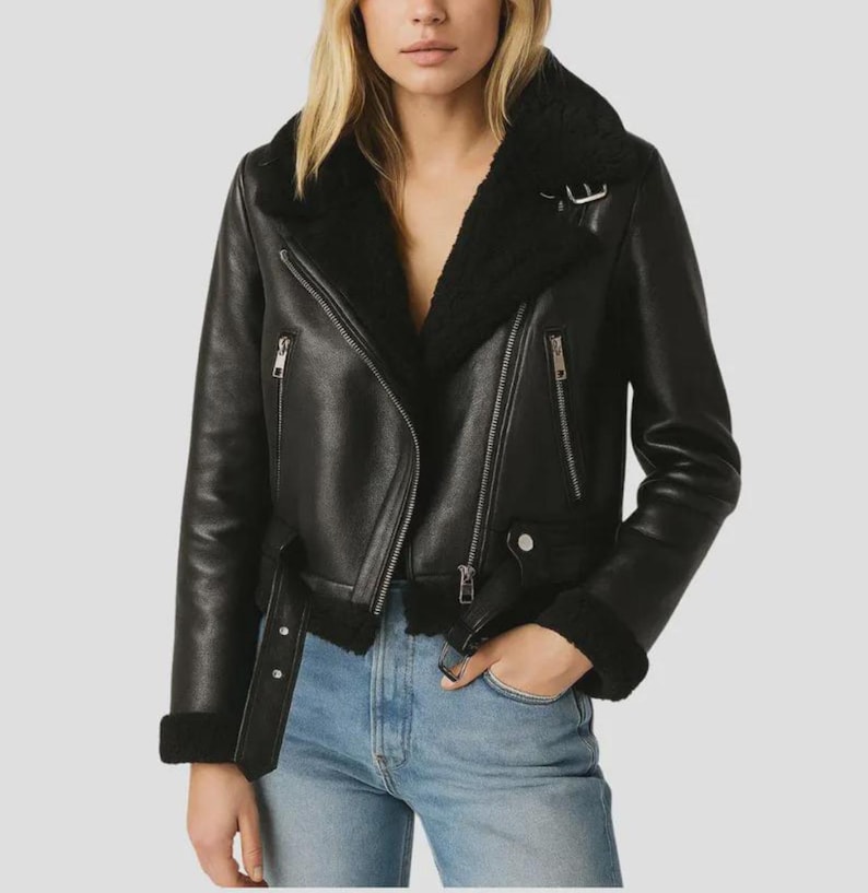 May include: Black leather jacket with a shearling collar and lining. The jacket features a classic asymmetrical zipper closure, multiple zippered pockets, and a belt detail at the waist. The sleeves have shearling cuffs. The jacket is worn over blue jeans.