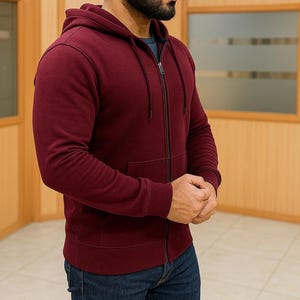 May include: A burgundy zip-up hoodie with a drawstring hood and a front pocket. The hoodie has a dark zipper and dark stitching details. The person is wearing blue jeans.