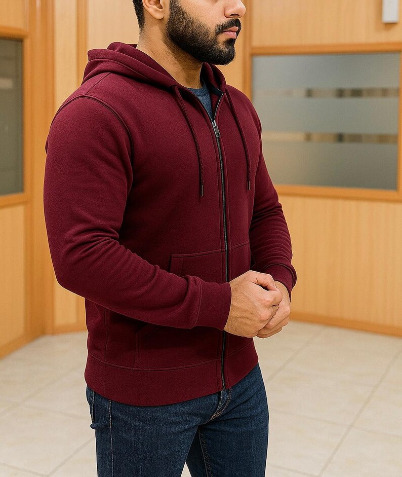 May include: A burgundy zip-up hoodie with a drawstring hood and a front pocket. The hoodie has a dark zipper and dark stitching details. The person is wearing blue jeans.