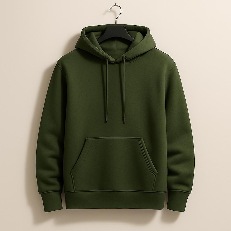K&ouml;nnte beinhalten: Ein einfarbiger olivgr&uuml;ner Kapuzenpullover mit Kordelzug und K&auml;ngurutasche. Der Pullover h&auml;ngt an einem schwarzen Kleiderb&uuml;gel vor einem neutralen Hintergrund. Es ist ein l&auml;ssiges Kleidungsst&uuml;ck.