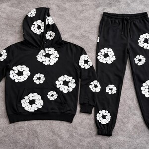 May include: Black hoodie and sweatpants set with white floral designs. The hoodie has a front pocket and hood. The sweatpants have a drawstring waist and cuffed ankles. The set is made of a soft material.