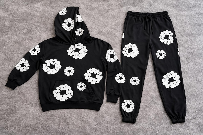 May include: Black hoodie and sweatpants set with white floral designs. The hoodie has a front pocket and hood. The sweatpants have a drawstring waist and cuffed ankles. The set is made of a soft material.