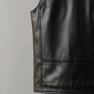 May include: Close-up of a black leather vest. The vest has a sleeveless design with a rounded neckline and a rectangular pocket detail at the bottom. The seams are accented with dark blue stitching.