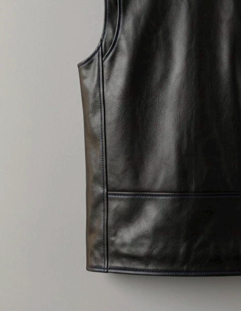 May include: Close-up of a black leather vest. The vest has a sleeveless design with a rounded neckline and a rectangular pocket detail at the bottom. The seams are accented with dark blue stitching.