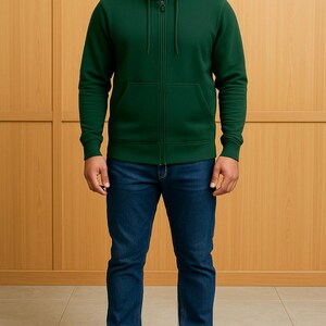 May include: A dark green zip-up hoodie with a front pocket and a hood. The hoodie is paired with blue jeans and black shoes. The background is a light brown wooden wall.