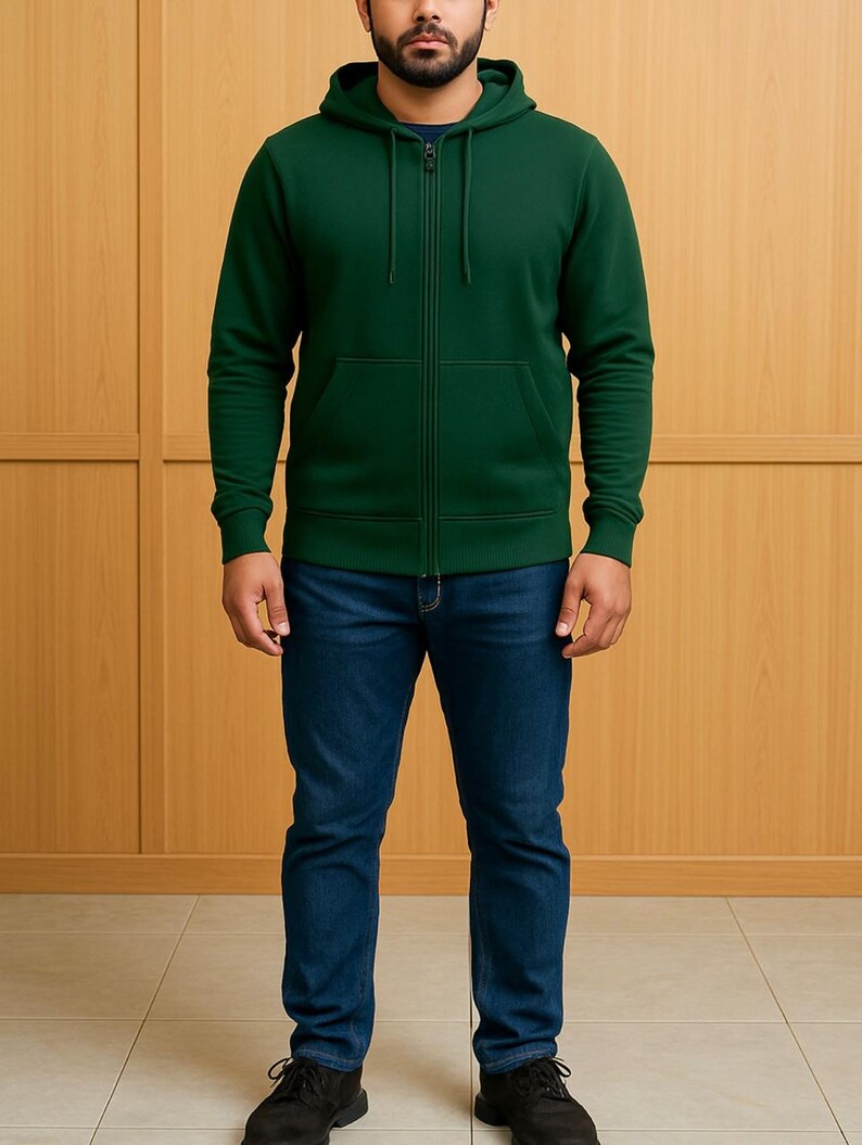 May include: A dark green zip-up hoodie with a front pocket and a hood. The hoodie is paired with blue jeans and black shoes. The background is a light brown wooden wall.