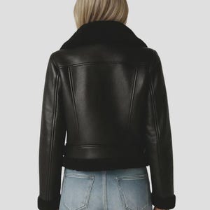 May include: Black faux leather jacket with a shearling collar and cuffs. The jacket features a classic aviator design with a belted waist and a zip closure. The jacket is shown from the back, with light blue jeans visible.